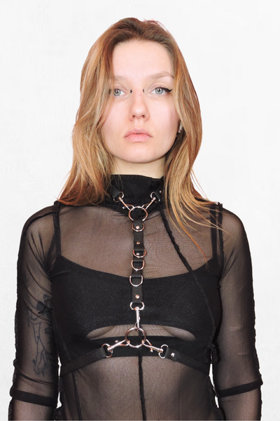 Model wearing a black harness that wraps around the neck like a choker, with an adjustable strap securing at the waist. Silver O-rings and hardware add a bold, edgy touch.