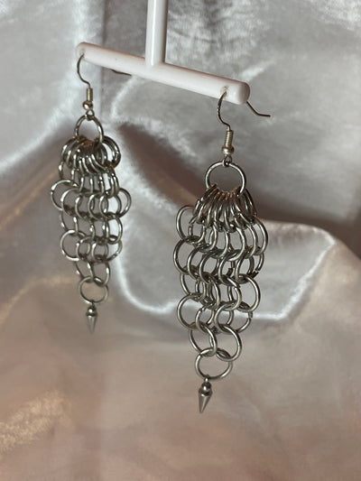 A close-up of chunky silver chain mail earrings with a bold, intricate weave. The earrings catch the light, showcasing their statement-making design against a minimalist background.