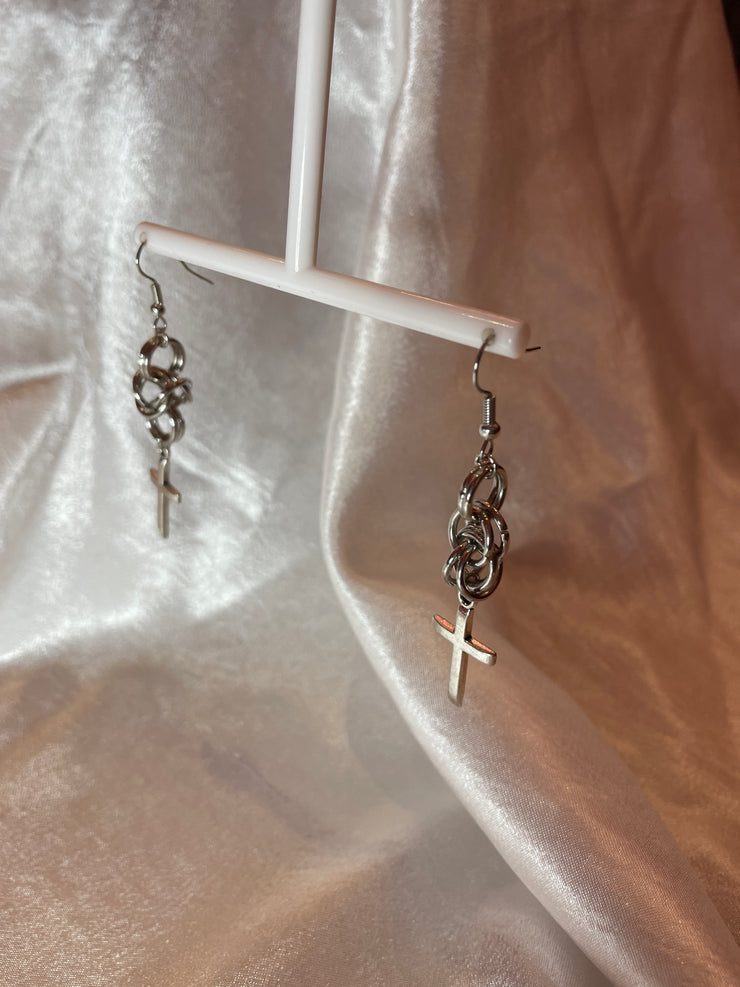 A close-up of elegant stainless steel chain mail earrings featuring a bold cross detail, hanging gracefully against a neutral backdrop. The intricate craftsmanship highlights their unique, handmade quality.